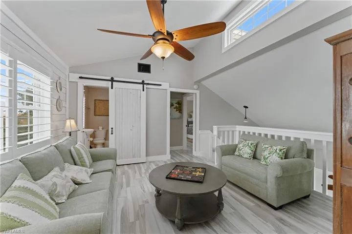 Vaulted ceiling with an overhead fan, large windows with plantation shutters, light wood-finish flooring, and a white barn door