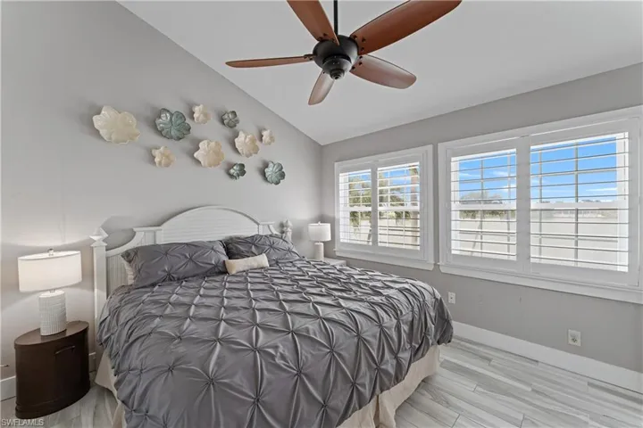 Vaulted ceiling bedroom featuring wood-finish flooring, two windows with plantation shutters, and a ceiling fan with wood-look blades