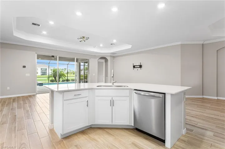 Kitchen with a raised ceiling, crown molding, light countertops, stainless steel dishwasher, and a center island with sink