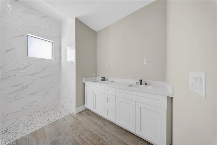 Spacious bathroom featuring a dual vanity with white cabinetry and light-toned countertops, a walk-in shower with patterned white tile walls and a mosaic tile floor, and wood-finish flooring
