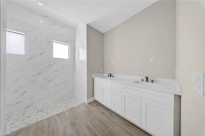 Spacious bathroom featuring a large shower with patterned tile walls and pebble flooring, dual vanity with white cabinetry, white countertops, and wood-finish flooring