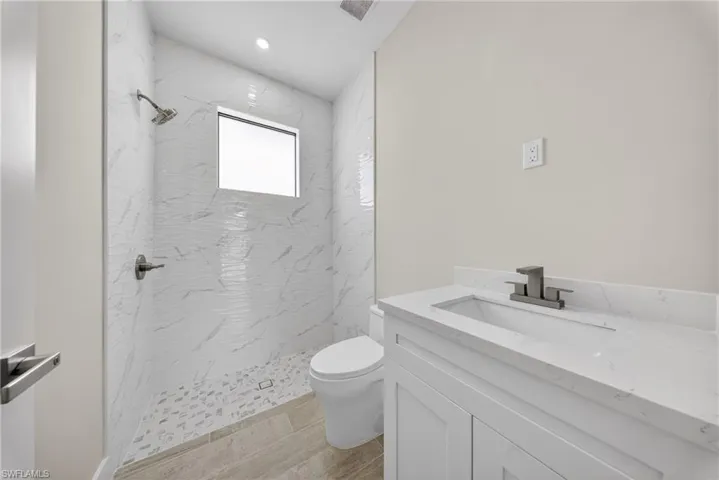 Bathroom featuring a walk-in shower with textured tile surround and mosaic floor, a single-pane window, a white vanity with integrated sink and stone countertop, and wood-finish flooring