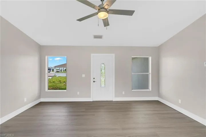 Bright interior space featuring wood-finish flooring, a white entry door with decorative glass, two windows, and a ceiling fan with integrated lighting