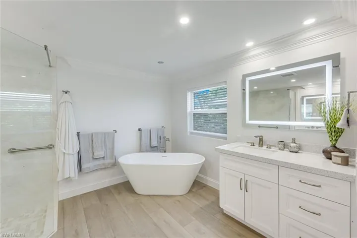 Primary Bathroom featuring a freestanding soaking tub, glass-enclosed shower, wood-finish flooring, a white vanity with a light-toned countertop, and an illuminated mirror