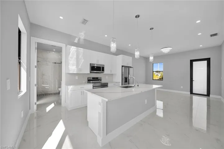 Kitchen featuring stainless steel appliances, white cabinets, an island with sink, hanging light fixtures, and light stone counters