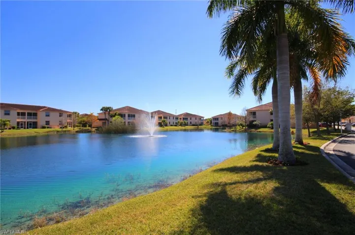 The community features a serene lake with a fountain, surrounded by lush green lawns and palm trees