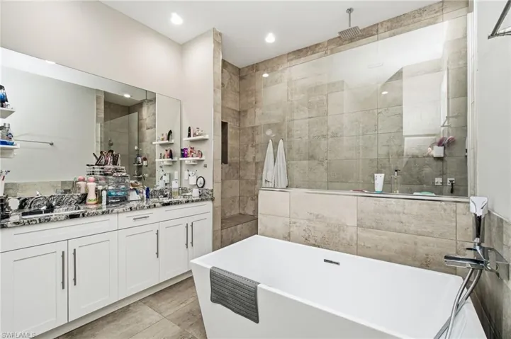 Freestanding soaking tub, walk-in shower with glass enclosure, dual vanity with white cabinetry, and large format tile flooring