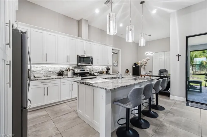 White shaker cabinetry with brushed nickel hardware, stainless steel appliances, granite countertops, tile backsplash, and three pendant lights above the island