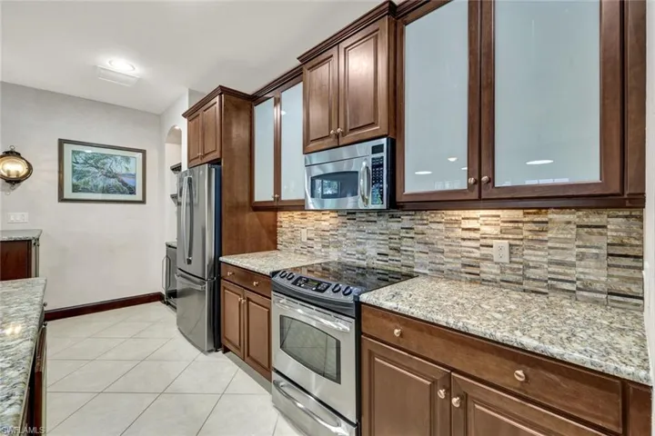 Kitchen featuring light stone counters, backsplash, appliances with stainless steel finishes, glass insert cabinets, and light tile patterned floors