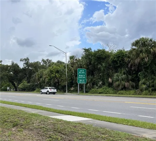 Roadside location featuring a paved multi-lane road, concrete sidewalk, and mature tropical foliage