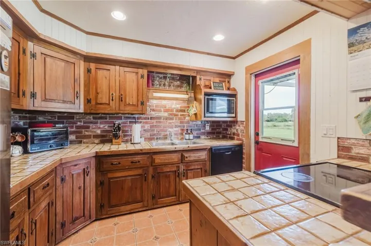 Wood cabinetry with black hardware, brick backsplash, tile countertops, integrated microwave, and a tile-top island with a built-in cooktop