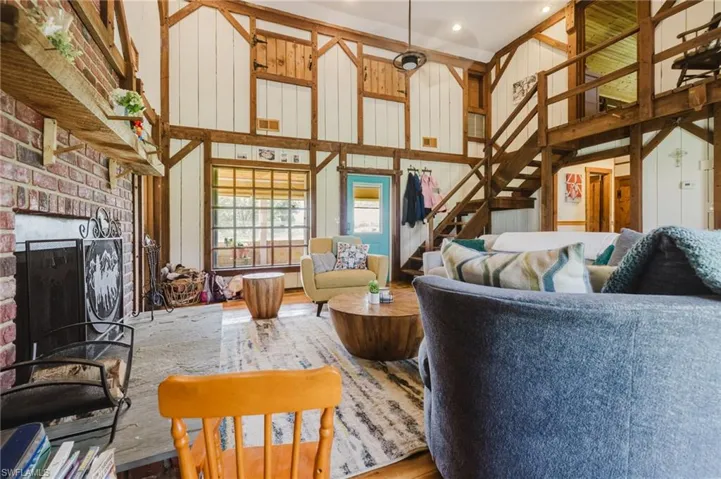 Exposed timber framing, brick fireplace with stone hearth, wood-finish flooring, two-story ceiling, and open staircase