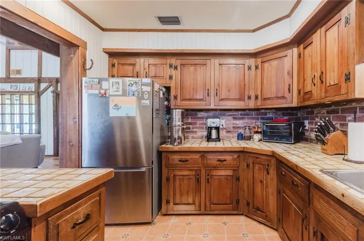 Wood-finish cabinetry with decorative hardware, tile countertops, brick-finish backsplash, and terracotta-style floor tiles