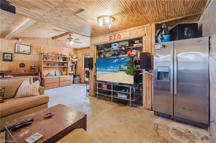 Vaulted wood-paneled ceiling with exposed beams, wood-plank walls, concrete flooring, built-in shelving, and a stainless steel refrigerator