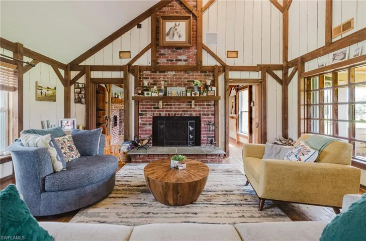 Vaulted ceiling with exposed wood beams, brick fireplace with stone hearth, wood-finish flooring, and large multi-pane windows