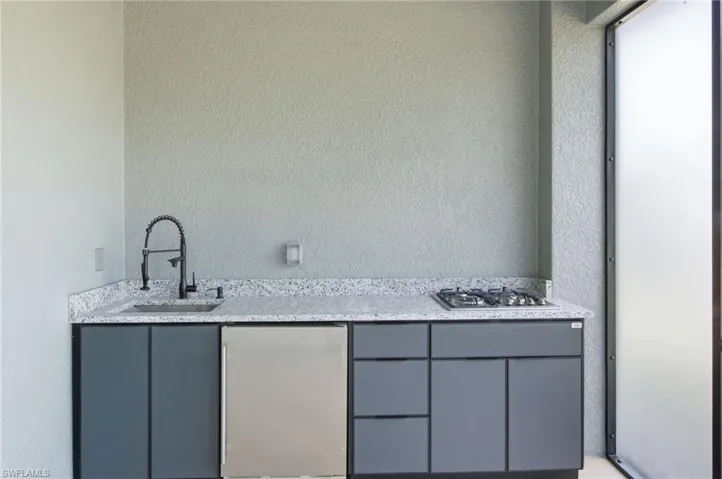 Indoor wet bar featuring light stone counters, stainless steel appliances, a textured wall, and gray cabinetry