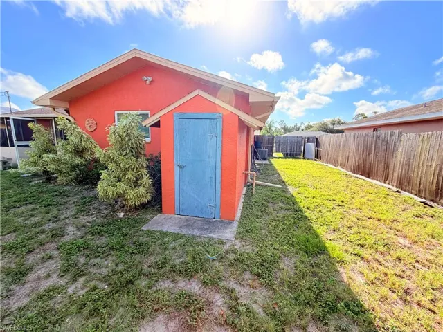 Exterior featuring stucco siding, an attached shed with a blue door, a partial screen enclosure, mature landscaping, and a wooden privacy fence