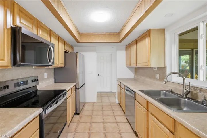 Galley kitchen featuring wood cabinetry, stainless steel appliances, a double basin sink with a high-arc faucet, a tray ceiling, and tiled flooring