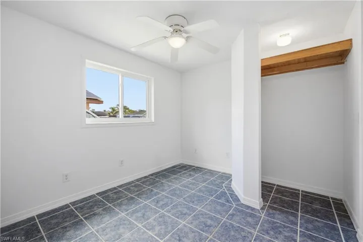 Room featuring blue tiled flooring, a window with white trim, a ceiling fan with integrated lighting, and a built-in closet area with an exposed wood beam shelf and ceiling light