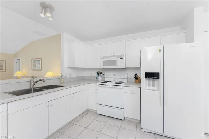 Kitchen with light tile patterned flooring, vaulted ceiling, sink, white cabinets, and white appliances