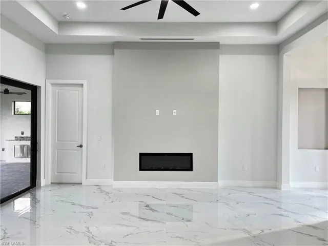 Unfurnished living room featuring ceiling fan, a glass covered fireplace, a tray ceiling, light marble finish flooring, and recessed lighting