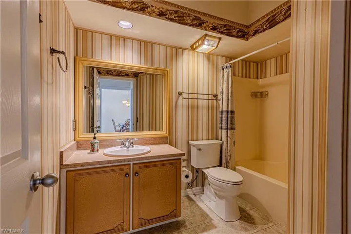 Guest bathroom w/ shower / bath combination with curtain, vanity, light tile patterned flooring, wallpapered walls & recessed lighting.