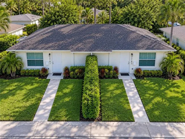 Duplex property featuring a shingled roof, white exterior, front entryways, and manicured landscaping with mature shrubs and palm trees