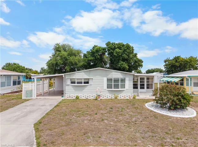 Light-colored siding exterior with a covered carport, white picket fence, and a paved driveway