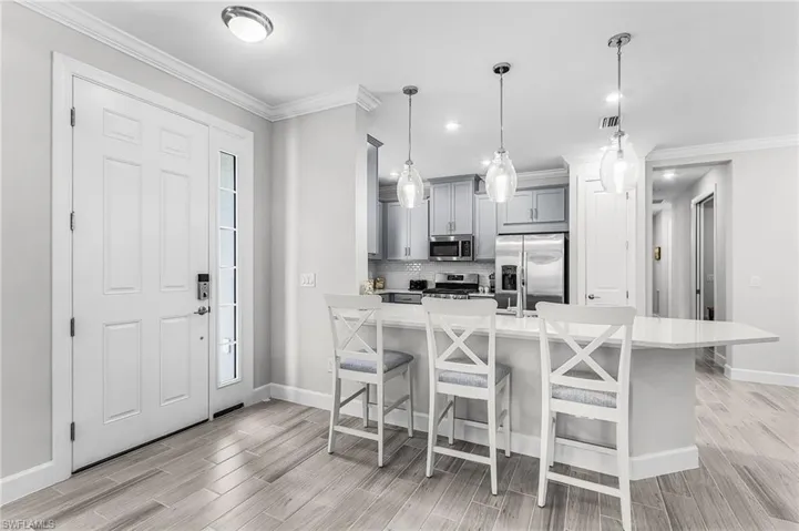 Open-concept kitchen and entryway featuring light wood-finish flooring, a white paneled entry door with sidelight, a kitchen island with seating, pendant lighting, and stainless steel appliances