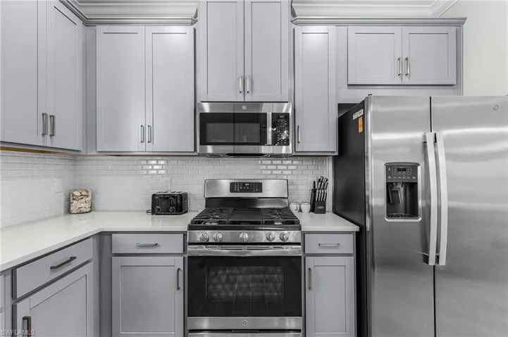 Kitchen featuring light gray shaker-style cabinetry, stainless steel appliances, white subway tile backsplash, white countertops, and crown molding accents