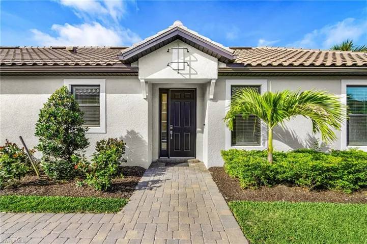 Tiled roof residence with a paver walkway, mature landscaping, stucco exterior, and dark paneled entry door