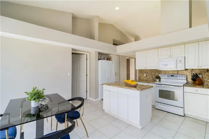 Kitchen with granite backsplash, white cabinets, high vaulted ceiling, white appliances, and a kitchen island