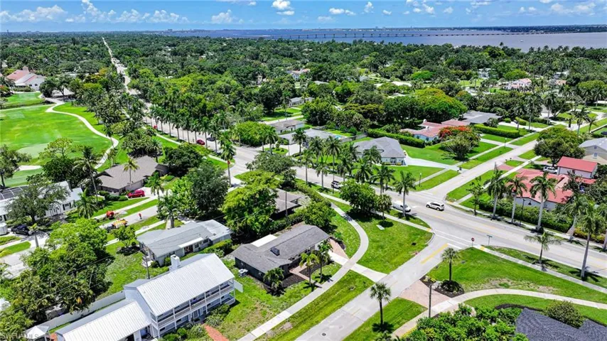 Aerial perspective highlighting a residential area with a prominent golf course, mature trees, and a distant body of water with a bridge structure