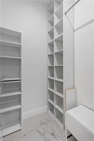 Walk-in closet featuring white built-in shelving, white baseboards, a chrome hanging rod, and light-toned tile flooring with subtle veining