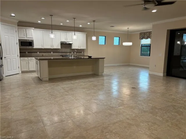 Kitchen featuring backsplash, stainless steel microwave, an island with sink, ceiling fan, and white cabinets