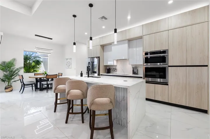 Two tone kitchen featuring hanging light fixtures, a kitchen island with sink, a breakfast bar area, stainless steel appliances, and light stone counters