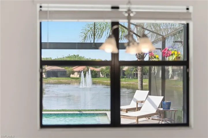Expansive window framing a screened lanai with a swimming pool, overlooking a lake with a fountain and tropical landscaping