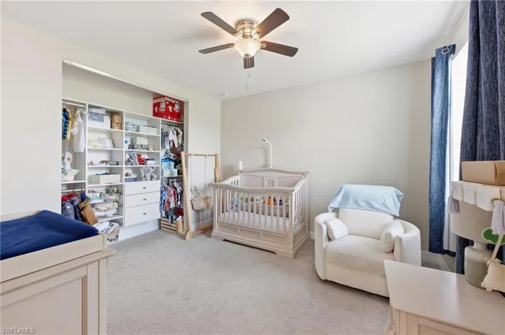 Bedroom featuring light-toned walls, gray carpeting, a built-in closet system with shelving and drawers, a window with drapery, and an overhead ceiling fan with lighting