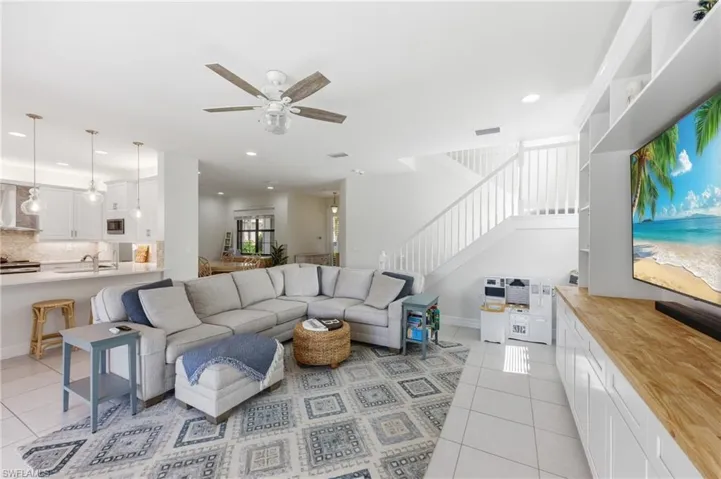 Open concept living area featuring a kitchen with white cabinetry and pendant lighting, a built-in media center with a wood-finish countertop, a staircase with white balusters, a ceiling fan, and white tile flooring