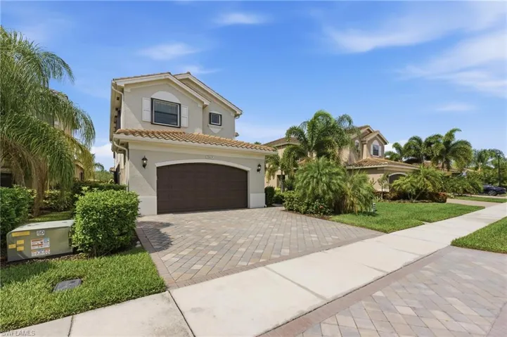 Two-story stucco residence featuring a tile roof, an arched entry garage, a paver driveway, mature landscaping, and an adjacent concrete sidewalk