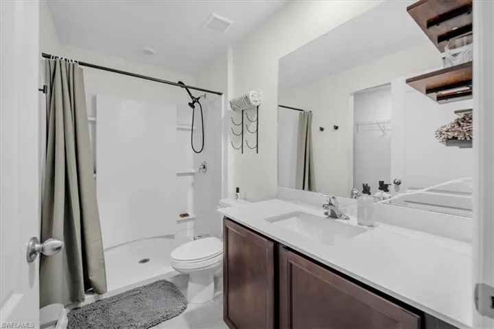 Bathroom featuring a white vanity with a composite countertop and integrated basin, full-width wall mirror, dark wood-finish cabinetry, and a shower stall with a matte black fixture