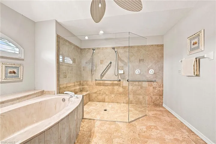 Spa-inspired primary bathroom featuring a frameless glass-enclosed walk-in shower with dual showerheads and body jets, heated floor, and travertine tile surround