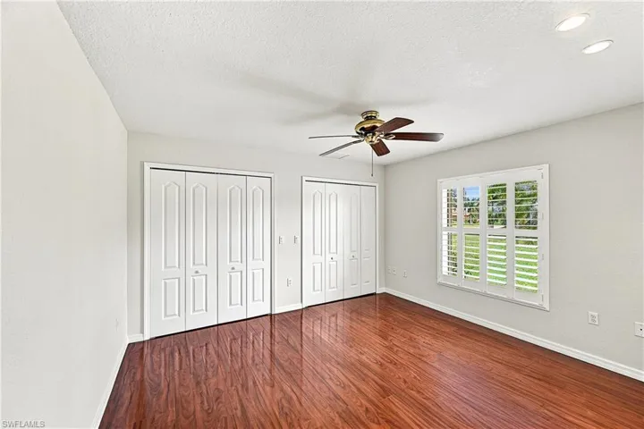 Spacious guest bedroom showcasing rich wood-finish flooring, white panel closet doors with multiple storage sections, white plantation shutters on expansive windows, ceiling fan, and bright recessed lighting