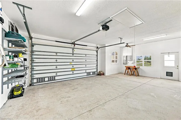 Spacious garage featuring a hurricane rated garage door, concrete flooring, fluorescent lighting, and built-in shelving, pull-down stairs to attic area. Windowed corner area with fan is workshop space, with electric panels pre-wired for portable generator.