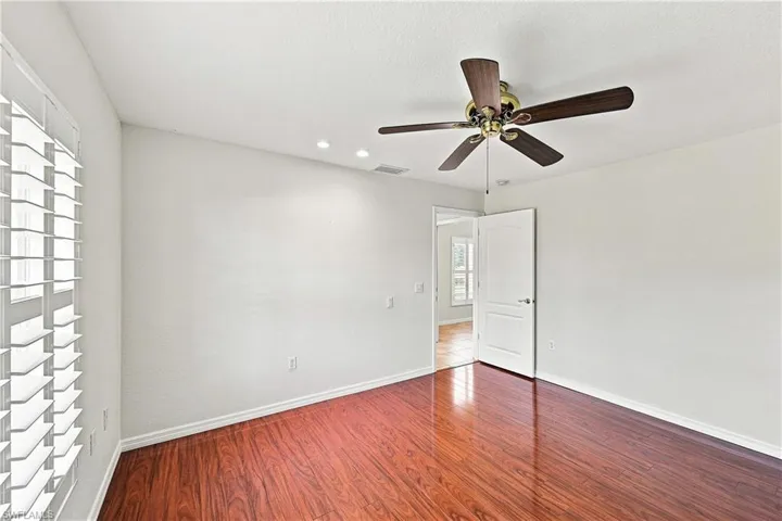 Spacious bedroom featuring rich wood-finish flooring, recessed ceiling lighting, ceiling fan with brushed metal and dark blade finish, and abundant natural light from shuttered windows