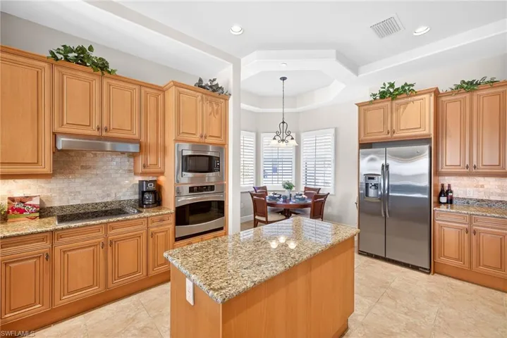 Kitchen featuring backsplash, appliances with stainless steel finishes, a tray ceiling, light stone countertops, and decorative light fixtures