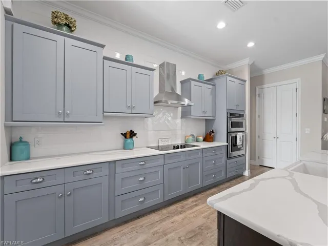 Kitchen with gray cabinets, crown molding, wall chimney range hood, light wood-type flooring, and tasteful backsplash