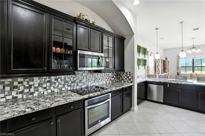 Kitchen featuring dark cabinetry, appliances with stainless steel finishes, glass insert cabinets, backsplash, and light stone countertops