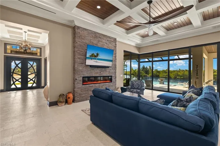 Living room featuring ornamental molding, coffered ceiling, ceiling fan with notable chandelier, beam ceiling, and a stone fireplace