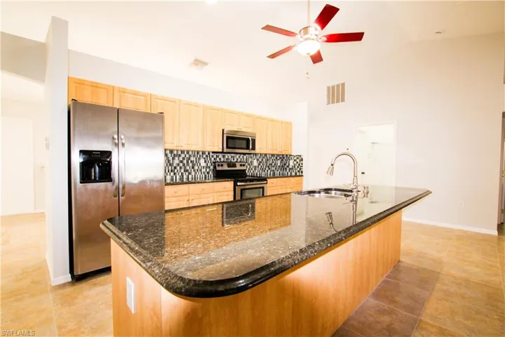 Kitchen with stainless steel appliances, dark stone counters, light brown cabinets, an island with sink, and lofted ceiling